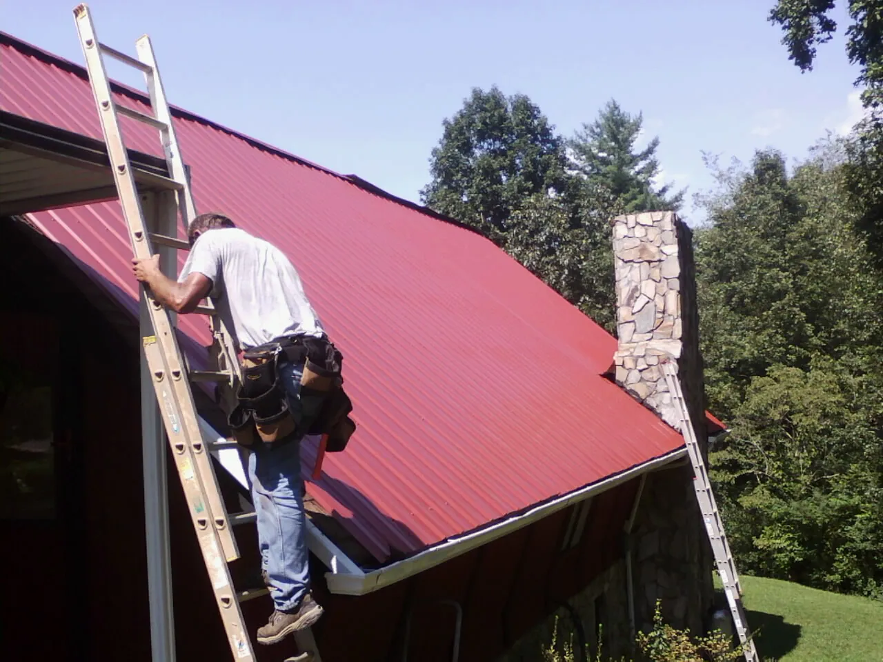 Roofer on a ladder beside a red metal roof and stone chimney — Soffit & Fascia Repair by Roofing Brothers in Coalfield